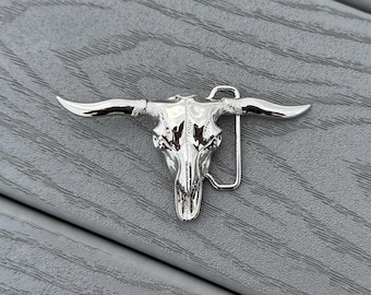 May include: A silver-colored belt buckle in the shape of a bull skull. The buckle has a detailed design of the skull's features, including horns and facial structure. The buckle is made of a shiny metal and has a standard belt loop.