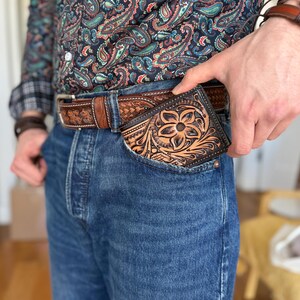 May include: A brown leather wallet with a floral design is tucked into the front pocket of a pair of blue jeans.