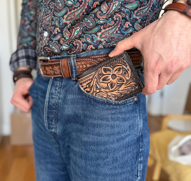 May include: A brown leather wallet with a floral design is tucked into the front pocket of a pair of blue jeans.