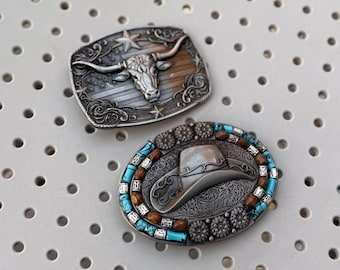 May include: Two silver-toned belt buckles. One rectangular buckle features a bull's head with longhorns and stars. The other oval buckle has a cowboy hat design, surrounded by turquoise and brown accents, and decorative metalwork.