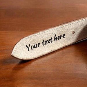 以下が含まれることがあります： 薄い茶色の革製のベルトのクローズアップ。ベルトには「Your text here」という文字が刻まれています。