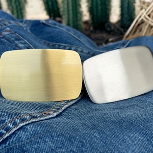 May include: Two metal belt buckles, one gold and one silver, on a blue denim background.
