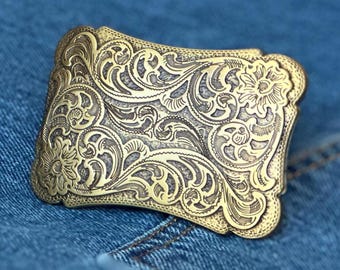 May include: A rectangular brass-toned belt buckle featuring an intricate, raised floral and scrollwork engraving across the entire surface. The metal has a polished finish on the raised patterns with a darkened, recessed background texture.