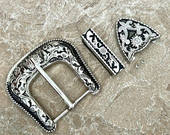 May include: Silver-toned western belt buckle set with intricate floral and sunburst designs. The set includes a buckle, belt loop, and tip, all with a detailed, engraved pattern. The pieces are arranged on a neutral, textured surface.