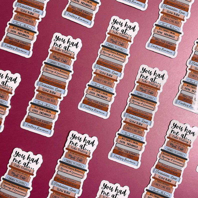 Op de afbeelding: Een stapel stickers met een ontwerp van een stapel boeken. Elke sticker heeft een andere boektitel, waaronder "Forbidden Love", "Slow Burn", "Only One Red", "Best Friend's Obsession", "Love Triangle", "Small Town Romance", "Insta Love" en "Fantasy Romance". De stickers hebben een lichtbruin en blauw kleurenschema. De tekst op de stickers luidt "You had me at... Fake Dating, Meet Cute".
