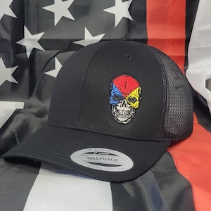 Puede incluir: Gorra de camionero negra de malla con una calavera negra bordada en la parte delantera. Las cuencas de los ojos de la calavera están rellenas de rojo, azul y amarillo. La gorra tiene una visera negra y una etiqueta negra y blanca que dice "The Authentic Yupoong Snapback Yupoong Since 1974."