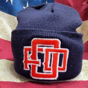May include: A navy blue knit beanie with a red and white embroidered letter "D" on the front.
