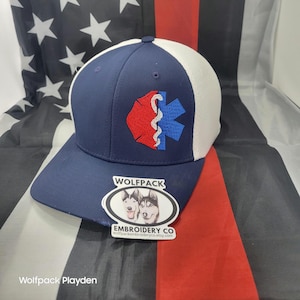 May include: A navy blue and white trucker hat with a red and blue medical symbol patch. The hat has a curved brim and a mesh back. A sticker with two husky dogs and the text "WOLFPACK EMBROIDERY CO" is on the brim.