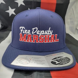 以下が含まれることがあります： 紺色のベースボールキャップに、白と赤の刺繍で「Fire Deputy Marshal」とロゴが入っています。キャップの側面にはFlexfit 110のラベルが付いています。