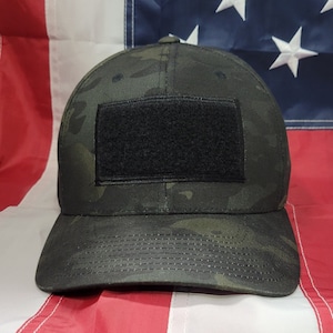 Puede incluir: Una gorra de béisbol de camuflaje negro y verde con un parche de velcro negro en la parte delantera. La gorra tiene una visera curvada y una corona estructurada.