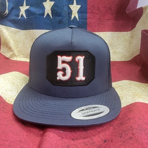 Op de afbeelding: Een donkerblauwe truckercap met een zwart en rood patch met het nummer "51" in wit. De pet heeft een mesh achterkant en een drukknopsluiting. De pet is gelabeld "The Authentic Yupoong Snapback Since 1974".