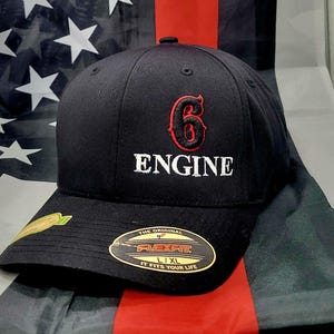 Peut inclure: Casquette de baseball noire avec un "6" rouge et "ENGINE" blanc brodés à l'avant. La casquette a une étiquette Flexfit noire avec le texte "The Original Flexfit L/XL It Fits Your Life".