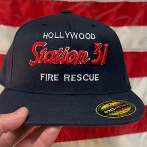 Op de afbeelding: Een marineblauwe baseballpet met witte stiksels en de tekst "Hollywood Station 31 Fire Rescue" geborduurd in rood en wit. De pet heeft een zwart-gele sticker aan de zijkant met de tekst "210 Fitted".