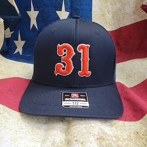 以下が含まれることがあります： 前面に赤と白の刺繍で「31」が施されたネイビーブルーのトラッカーハット。メッシュバックと、黒い調節可能なストラップに「リチャードソン」のラベルが付いています。