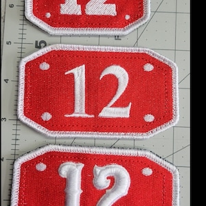 Op de afbeelding: Drie rode stoffen patches met witte stiksels en witte cijfers "12" in verschillende stijlen. De patches zijn rechthoekig met afgeronde hoeken.