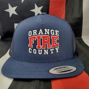 Peut inclure: Casquette de camionneur bleu marine avec les mots "ORANGE FIRE COUNTY" brodés en blanc et rouge. Le chapeau a un dos en filet et une visière plate. Une étiquette argentée et noire est sur la visière.
