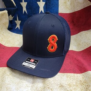 May include: A navy blue baseball cap with a red and orange number 8 embroidered on the front. The cap is a trucker style with a mesh back and a black adjustable strap. The cap has a red and white label with the text "Richardson. Original Trucker 112 Adjustable."