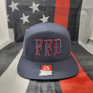 Op de afbeelding: Een donkerblauwe trucker-pet met een platte klep en rode geborduurde letters die "FFD" zeggen. De pet ligt op een Amerikaanse vlag.