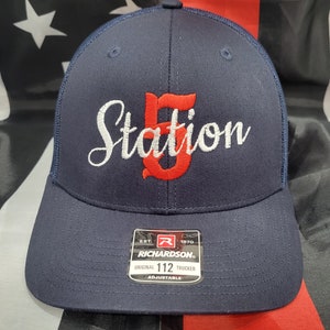 Può includere: Un cappello da camionista blu navy con una rete bianca sul retro. La parte anteriore del cappello ha un logo ricamato rosso e bianco che dice "Station 5". Il cappello ha anche un'etichetta in bianco e nero che dice "Richardson" e "Original 112 Trucker Adjustable".