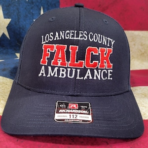 May include: A navy blue trucker hat with white mesh back. The hat has white embroidery that reads "Los Angeles County Falck Ambulance" with the word "Falck" in red. The hat has a black and red Richardson label with the text "Original 112 Trucker Adjustable".
