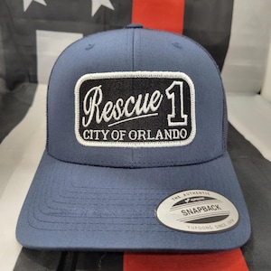 May include: A navy blue trucker hat with a white mesh back. The hat has a black and white patch that says "Rescue 1 City of Orlando". The hat also has a white "Snapback" label on the back.