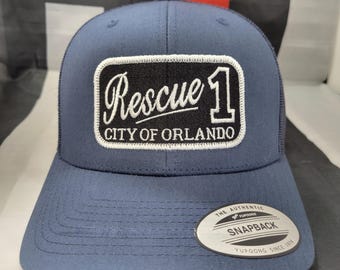 Könnte beinhalten: Eine marineblaue Trucker-Kappe mit weißem Netzrücken. Die Kappe hat einen schwarz-weißen Aufnäher mit der Aufschrift "Rescue 1 City of Orlando". Die Kappe hat auch ein weißes "Snapback"-Etikett auf der Rückseite.