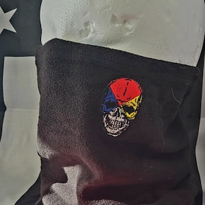 May include: Black fleece neck gaiter with a colorful skull design. The skull is divided into red, blue, and yellow sections. The background includes an American flag with a red stripe.