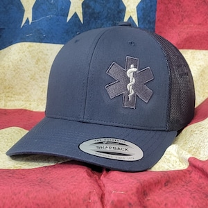 Puede incluir: Una gorra de camionero azul marino y negra con un símbolo médico bordado en blanco y el texto "The Authentic Yupoong Snapback Yupoong Since 1974".