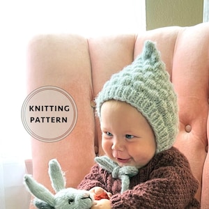 May include: A baby wearing a gray knitted hat sits in a pink armchair with a gray knitted bunny, a gray knitted pig, and a brown and white knitted panda.