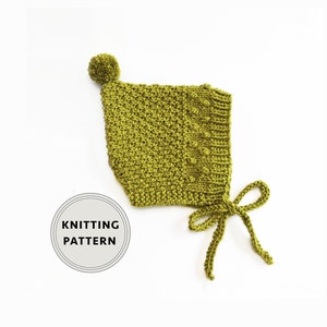 Op de afbeelding: Een groene gebreide baby muts met een pompon bovenop en strikjes aan de onderkant. De muts heeft een textuur in de steek en een klein knoopdetail aan de zijkant. Breipatroon.