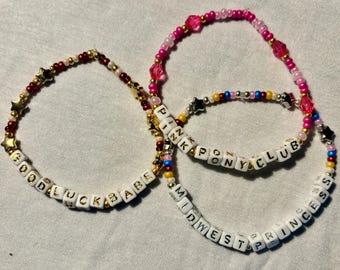 May include: Three beaded bracelets with white letter beads spelling out "Good Luck Babe", "Pink Pony Club", and "Midwest Princess". The bracelets are made with colorful beads in shades of pink, red, blue, and gold.