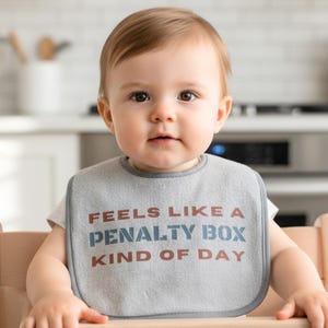 May include: A gray baby bib with the text "FEELS LIKE A PENALTY BOX KIND OF DAY" in brown and blue lettering. The bib is around the neck of a baby sitting in a wooden high chair.