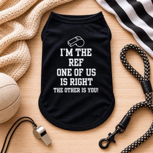 May include: Black dog t-shirt with white text that reads "I'M THE REF ONE OF US IS RIGHT THE OTHER IS YOU!" A silver whistle and a black and white striped leash are also visible.