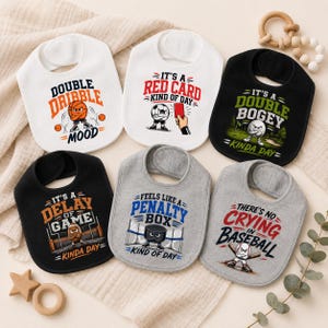 May include: Six baby bibs with sports-themed designs. The bibs are white, black, and grey, each with a different graphic and text related to sports such as basketball, soccer, golf, hockey, and baseball. They have a curved shape and a neck opening.
