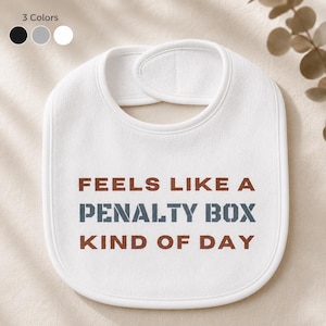 Op de afbeelding: Witte baby slabbetje met de tekst "FEELS LIKE A PENALTY BOX KIND OF DAY" in bruine en blauwe letters. De slab heeft een afgeronde vorm en een nek sluiting. Drie kleuropties worden getoond: zwart, grijs en wit.