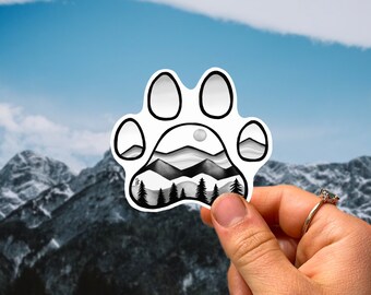 Puede incluir: Una pegatina de huella de pata en blanco y negro con una cordillera y árboles en su interior. La pegatina se sostiene frente a una cordillera.