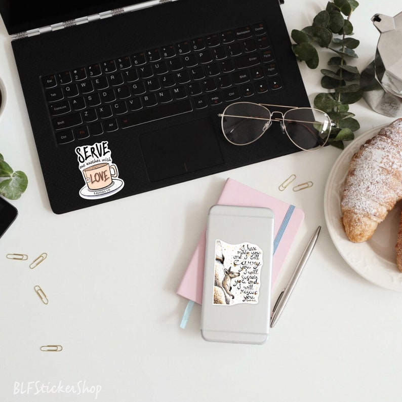 May include: A white laptop with a sticker that says "Serve one another with love" and a phone with a sticker that says "I have made you and I will carry you. I will sustain you and I will rescue you."