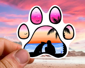 May include: A paw print sticker with a sunset scene inside. The sunset is pink, orange, and yellow with a silhouette of a person and a dog kissing. The paw print is white with a black outline.