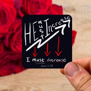 以下が含まれることがあります： 白と赤の文字が入った黒い正方形のステッカー。「HE must Increase」というテキストと上向きの矢印、「I must decrease」というテキストと下向きの矢印。聖書のヨハネ3:30も含まれています。