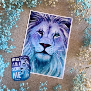 Op de afbeelding: Een print en sticker met een majestueuze leeuw met een blauwe en paarse manen en doordringende ogen. De sticker luidt "HERE AM I SEND ME" met het bijbelvers Jesaja 6:8.