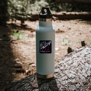 以下が含まれることがあります： 黒い蓋と金属製のベースが付いた、薄い青色のウォーターボトル。 ボトルには、矢印付きの「He must increase, I must decrease」と書かれた正方形のステッカーが付いています。 ボトルは森の中の丸太の上に置かれています。