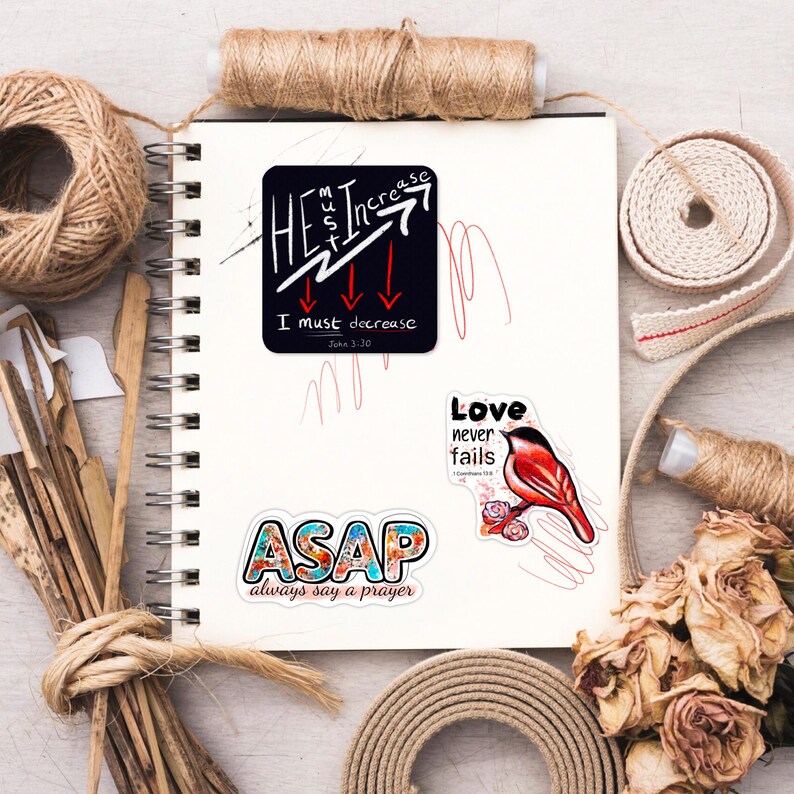 以下が含まれることがあります： 白いスパイラルノートに貼られたステッカーのコレクション。ステッカーには、「彼は増大しなければならない、私は減少しなければならない」、「ASAP always say a prayer」、「愛は決して失敗しない」などのテキストが含まれています。ステッカーはさまざまな色とデザインです。