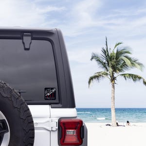 以下が含まれることがあります： 車の窓に貼られた、"HEAT"の文字と上向きと下向きの矢印が描かれた白黒のステッカー。ステッカーには「I May Persist」の文字も含まれています。背景には、ヤシの木と海のあるビーチが写っています。