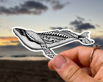 May include: A black and white patterned whale sticker with intricate details. The sticker is held in a hand against a blurred background of a sunset over the ocean.