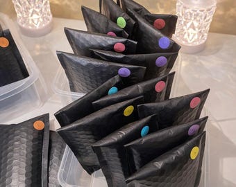 May include: A collection of black bubble mailers arranged in clear plastic containers. Each mailer features a small, circular colored sticker with the letter S, L, or E printed on it. The stickers are in shades of green, pink, purple, blue, and yellow.