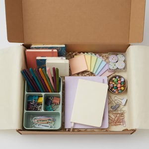 May include: An open cardboard box filled with stationery supplies. Includes notebooks, pens, sticky notes, paper clips, and decorative beads. The color palette is pastel with pops of color from the pens and beads.