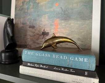 Könnte beinhalten: Eine Delfinfigur aus Messing auf einem Stapel von drei Büchern. Das oberste Buch ist blau und hat den Titel "The Glass Bead Game" auf dem Einband gedruckt. Das mittlere Buch ist schwarz und hat den Titel "Love in Another Town" auf dem Einband gedruckt. Das unterste Buch ist weiß und hat den Titel "Color of the Wind" auf dem Einband gedruckt.