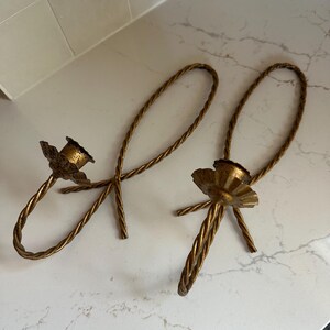 K&ouml;nnte beinhalten: Ein Paar goldene Kerzenhalter aus Metall mit einem gedrehten Seil-Design. Die Kerzenhalter haben die Form einer Schleife mit einem dekorativen Sockel.