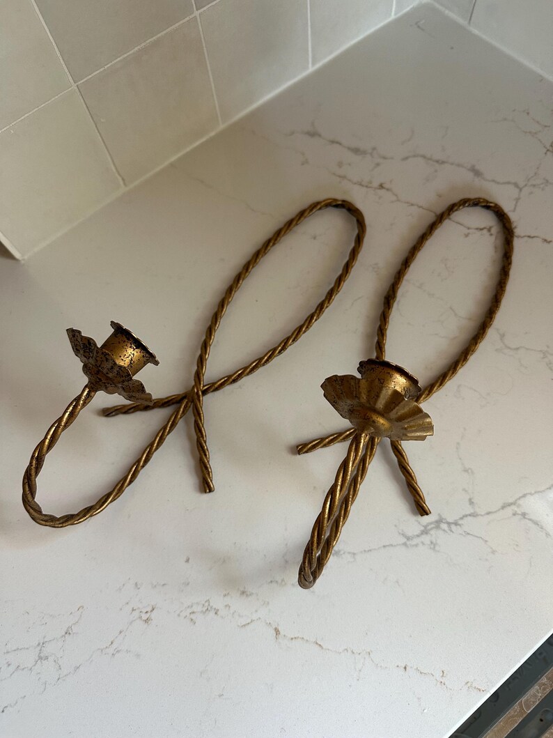 K&ouml;nnte beinhalten: Ein Paar goldene Kerzenhalter aus Metall mit einem gedrehten Seil-Design. Die Kerzenhalter haben die Form einer Schleife mit einem dekorativen Sockel.