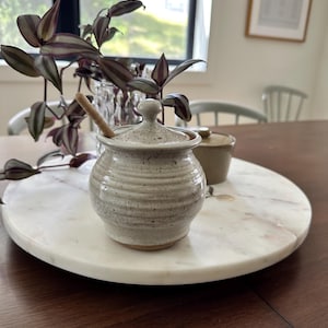 Puede incluir: Un pequeño tarro de miel de cerámica color hueso, de forma redonda, con acabado moteado y textura acanalada horizontal. Incluye una tapa a juego con pomo y un cucharón de madera para miel en su interior. Se aprecia la base de arcilla marrón.